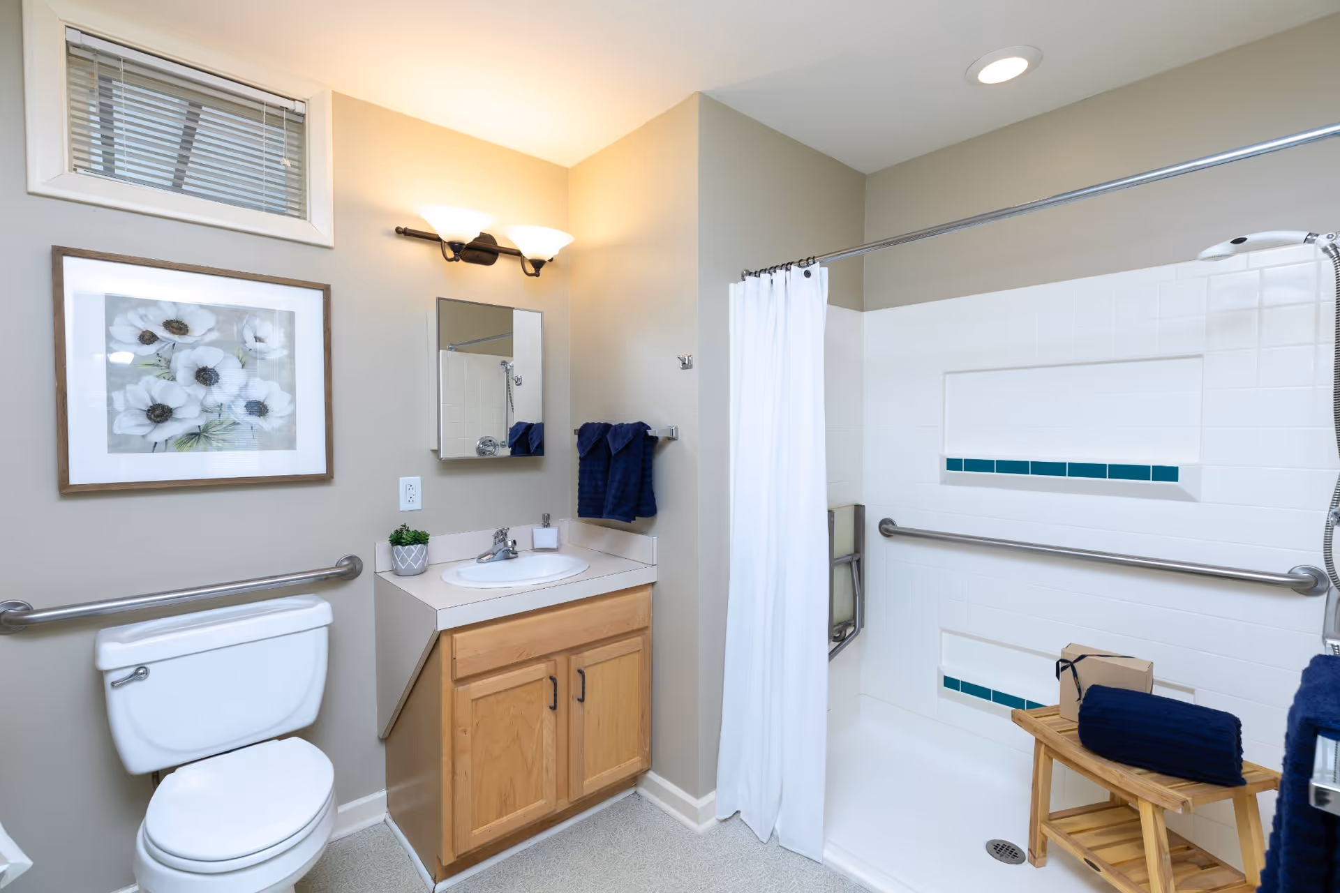 A clean and well-lit bathroom featuring a white toilet with a safety grab bar, a wooden vanity with a sink and a small plant, a mirror above the sink, a framed floral artwork on the wall, and a walk-in shower with a white curtain, grab bars, and a wooden stool holding a folded navy towel and a small box.
