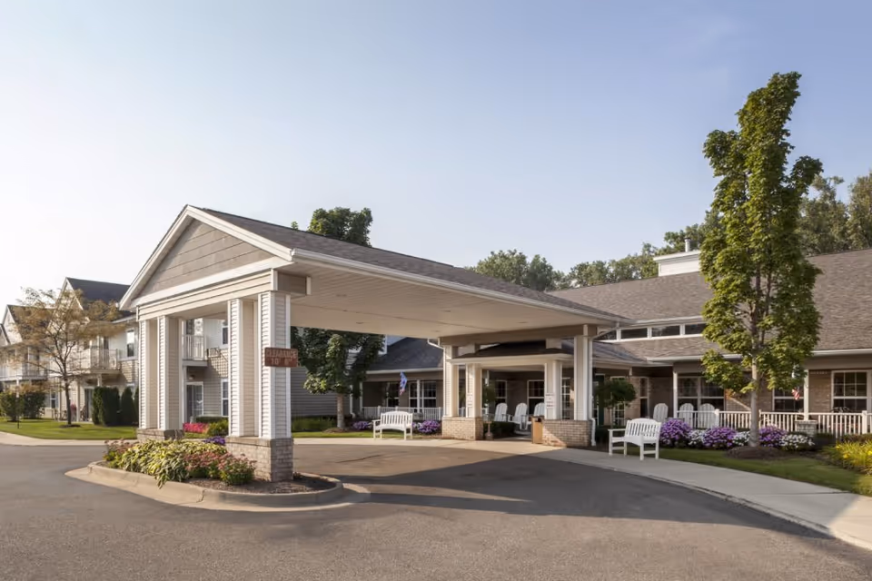 Exterior view of Pine Ridge of Plumbrook senior living facility entrance with a covered drop-off area, white benches, landscaped flower beds, and trees under a clear sky.