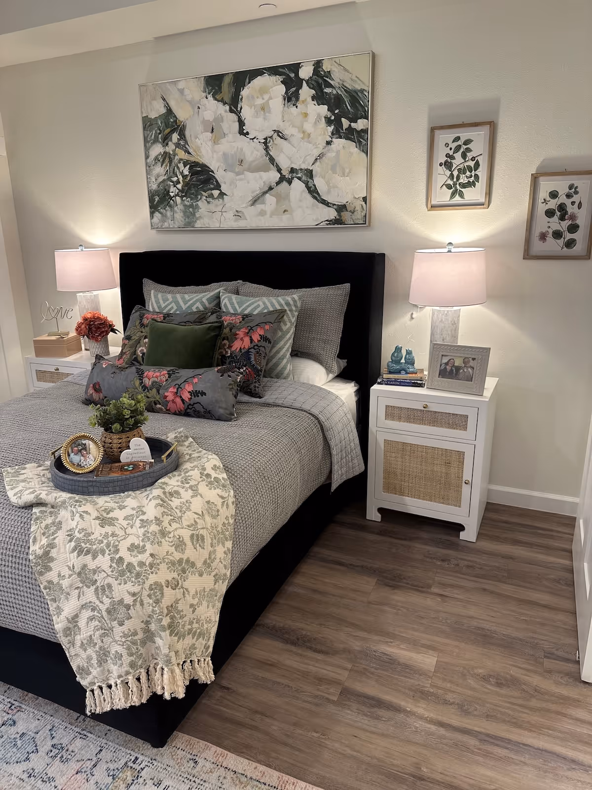 A cozy bedroom with a neatly made bed featuring multiple decorative pillows and a textured gray blanket. A floral-patterned throw is draped over the foot of the bed with a tray holding a small plant and framed photos. Two white nightstands flank the bed, each with a lamp and decorative items. Above the bed hangs a large floral painting, and two smaller botanical prints are on the adjacent wall. The room has wood flooring and a soft area rug partially visible.