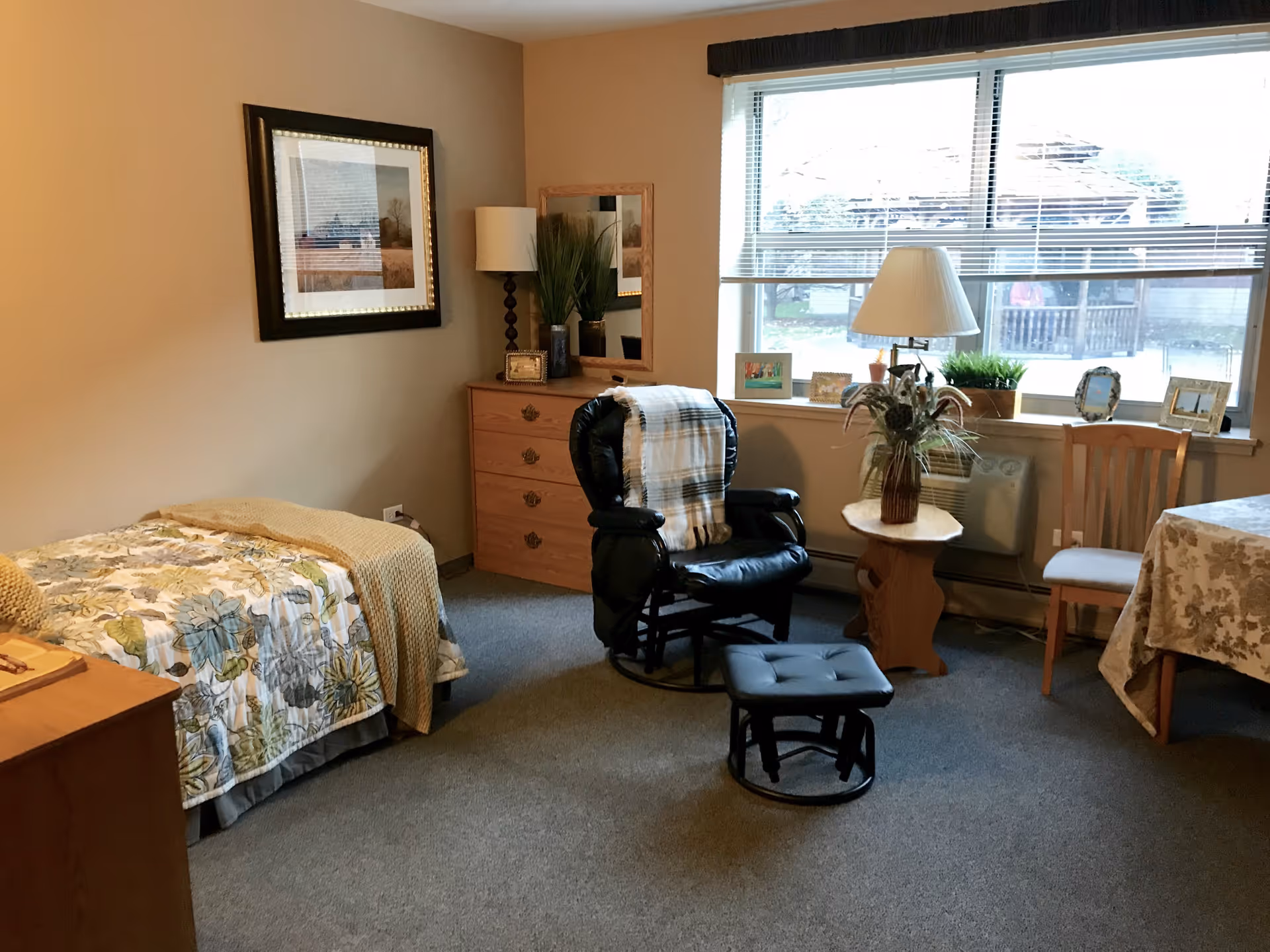 A cozy senior living bedroom with a single bed covered in a floral bedspread and a yellow blanket. There is a wooden dresser with a lamp and decorative items, a black leather rocking chair with a matching footrest and a plaid throw, a small wooden side table with a lamp and a vase of flowers, and a wooden chair next to a table covered with a floral tablecloth. A large window with blinds lets in natural light, and framed pictures are displayed on the windowsill and wall.