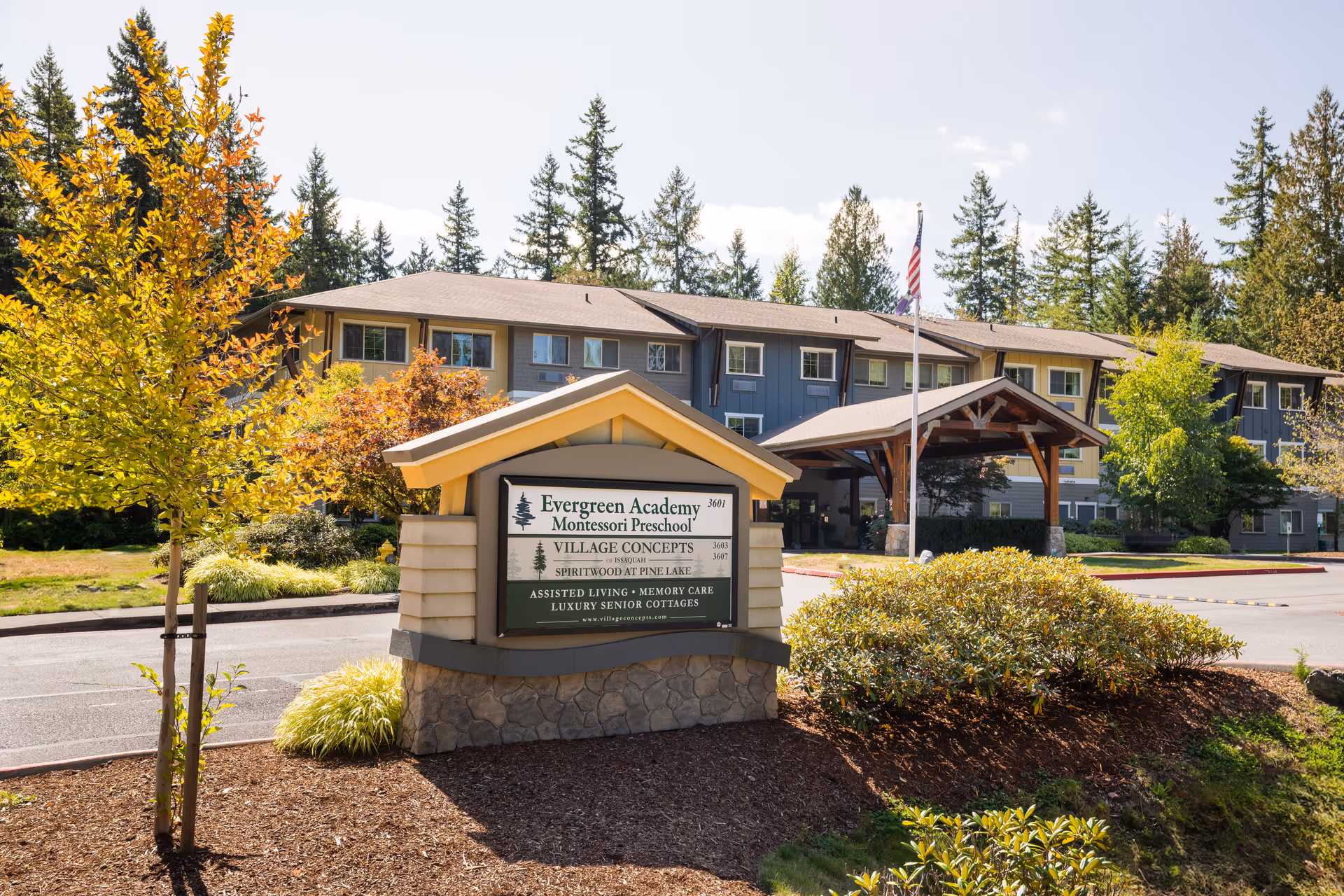 Exterior view of a senior living facility named Village Concepts of Issaquah-Spiritwood at Pine Lake, showing a multi-story building with a covered entrance, surrounded by trees and landscaping under a clear sky. A sign in front displays information about Evergreen Academy Montessori Preschool and the senior living services offered.