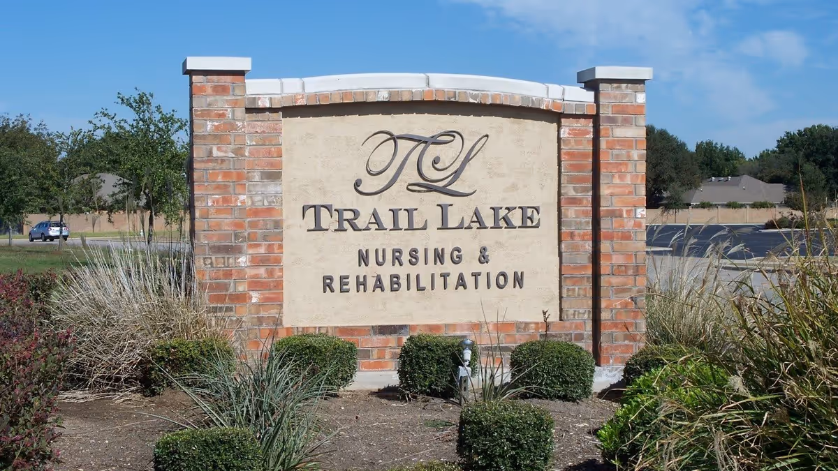 A brick and stucco sign for Trail Lake Nursing & Rehabilitation surrounded by landscaping with bushes and ornamental grasses, with a clear blue sky and some trees and houses in the background.
