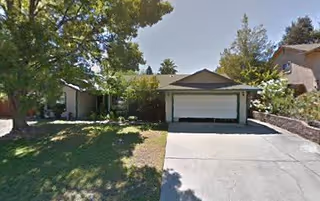 Front exterior view of a single-story house with a two-car garage, a driveway, and a lawn with trees and shrubs under a clear sky.