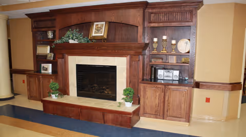 Interior view of a common area featuring a large wooden built-in shelving unit with a central fireplace. The shelves hold decorative items including framed pictures, plants, candles, and a stereo system. The floor has a polished, multicolored surface and the walls are painted in warm tones.