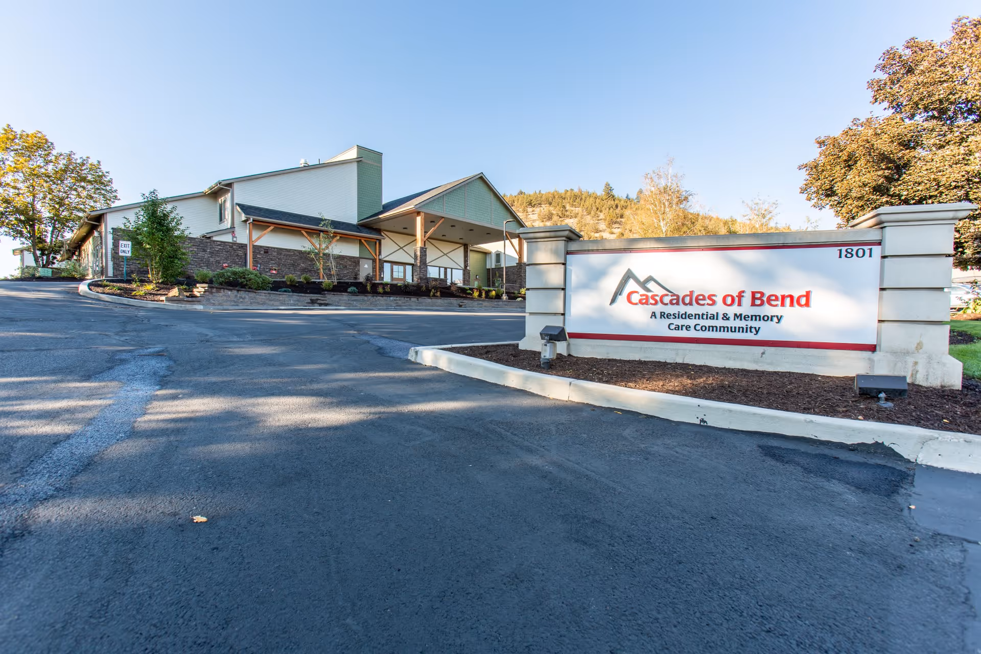 Exterior view of Cascades of Bend Retirement Community building with a large sign in front displaying the community name and description as a residential and memory care community. The building is surrounded by trees and landscaping under a clear blue sky.