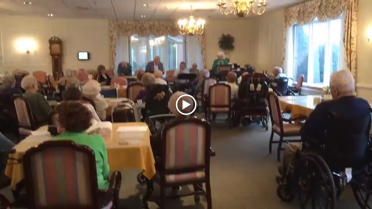 A group of elderly people seated in a large room with tables and chairs, some in wheelchairs, facing a speaker or performer at the front. The room has chandeliers, large windows with floral curtains, and a grandfather clock on the left wall.