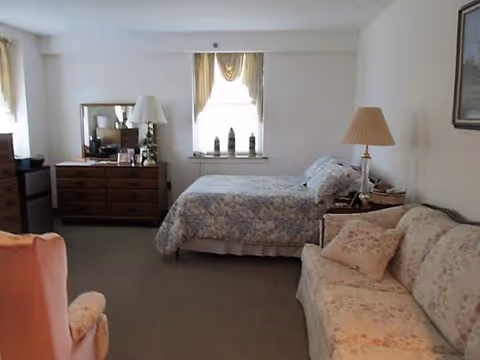 A cozy bedroom with a bed covered in floral bedding, a wooden dresser with a mirror, a table lamp, a floral-patterned sofa, and an armchair. There is a window with curtains letting in natural light.
