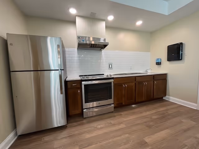 A modern kitchen area featuring a stainless steel refrigerator, an electric stove with oven, a stainless steel range hood, wooden cabinets with silver handles, a white subway tile backsplash, and a wall-mounted paper towel dispenser. The floor is covered with light wood laminate, and the walls are painted a light beige color.