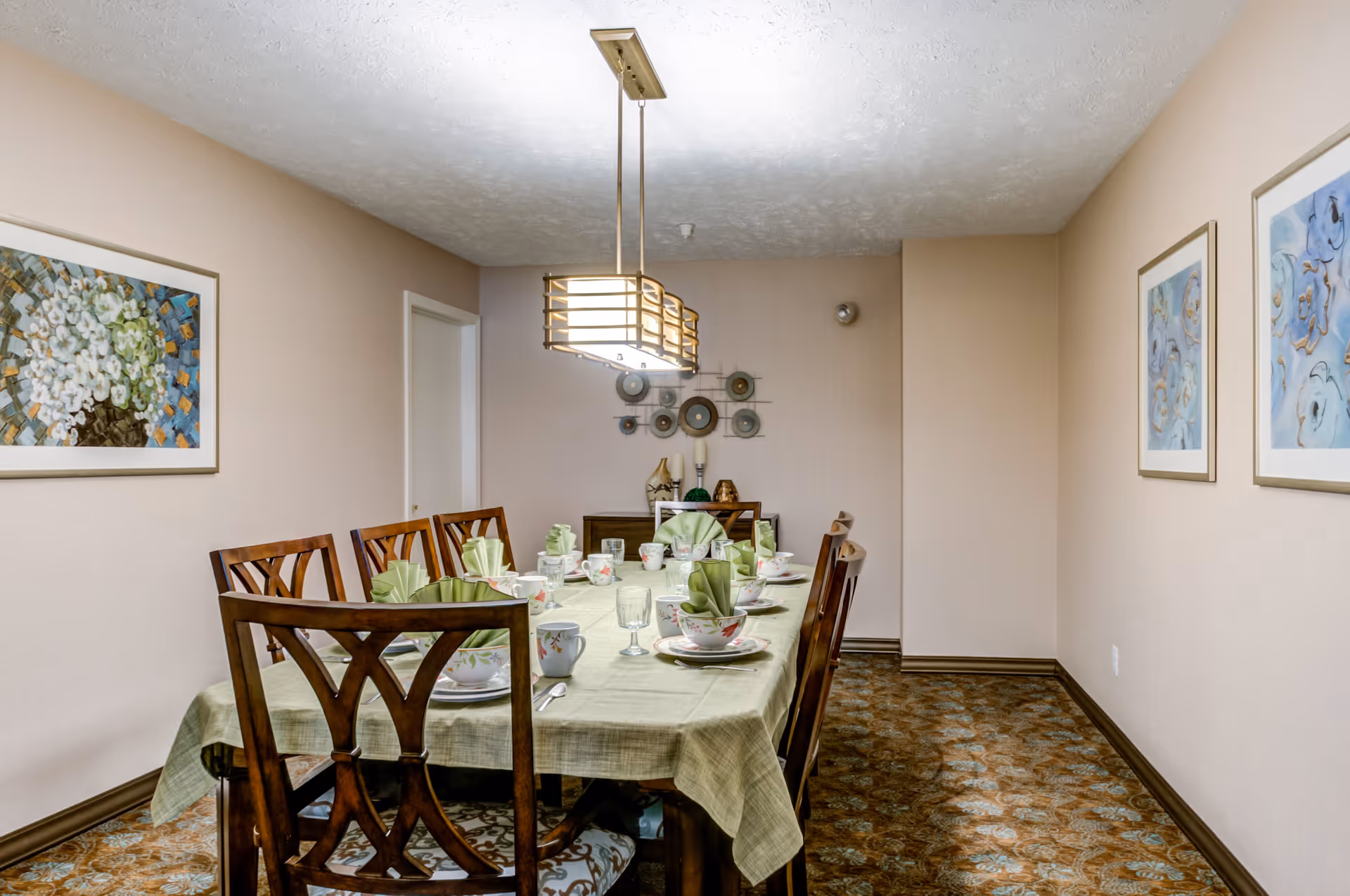 A dining room with a rectangular table covered with a light green tablecloth, set with floral-patterned dishes, cups, and folded green napkins. Six wooden chairs surround the table. The room has beige walls adorned with framed floral artwork and a decorative wall piece at the far end. A modern rectangular ceiling light fixture hangs above the table. The floor is covered with a patterned carpet.