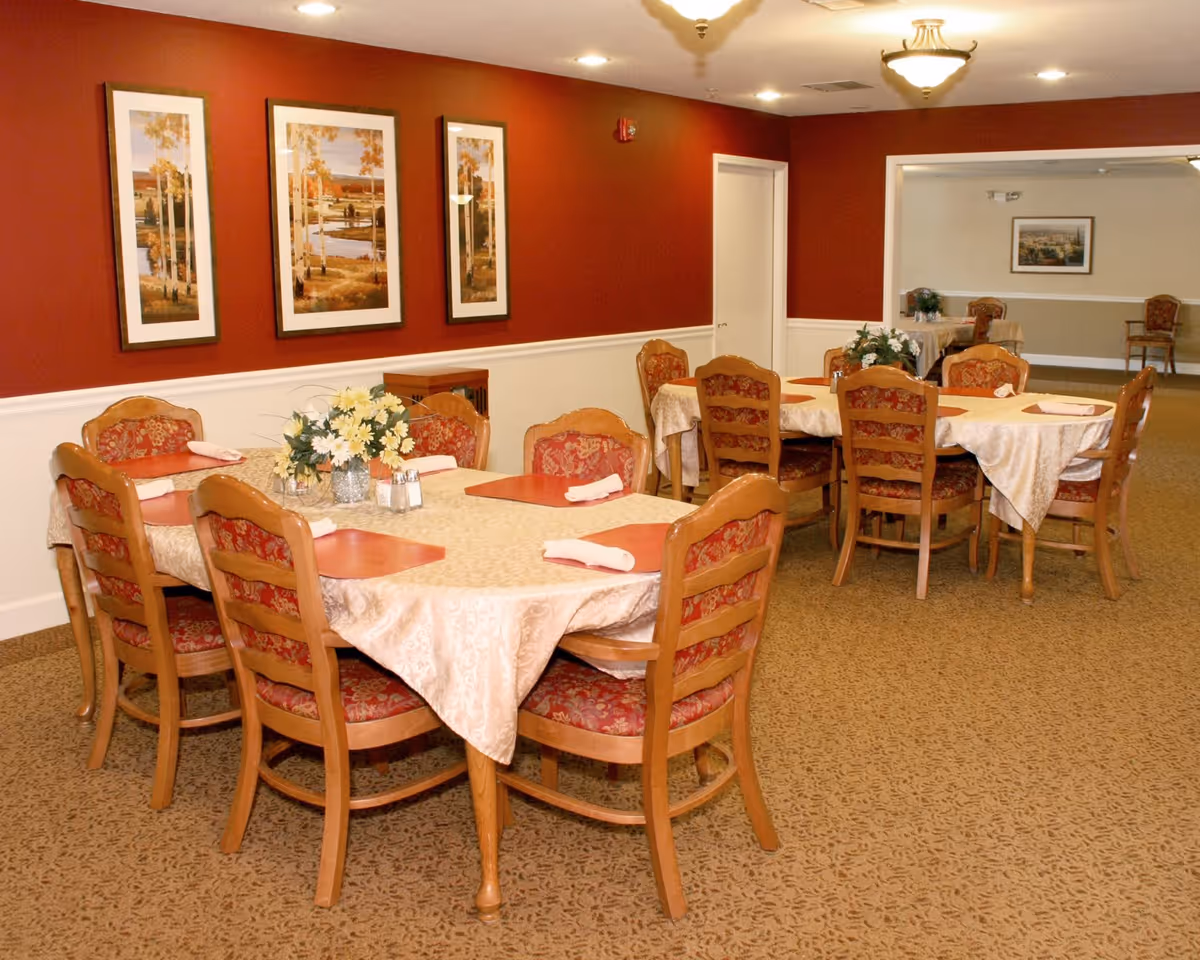 Dining room with two tables covered in beige tablecloths, each surrounded by wooden chairs with red patterned cushions. The tables have red placemats, white napkins, and floral centerpieces. The walls are painted red with white wainscoting and decorated with framed landscape paintings. The carpet is patterned in a neutral tone, and ceiling lights illuminate the room.