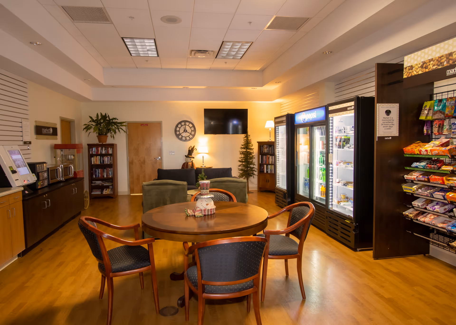 A cozy common area with a round wooden table surrounded by four chairs in the foreground. Behind the table, there are two green armchairs and a dark sofa facing a wall-mounted TV. To the right, there are refrigerated beverage coolers and a snack display rack. On the left side, there is a counter with a popcorn machine, microwave, and a touchscreen kiosk. The room has wooden flooring, a potted plant, bookshelves, and warm lighting.