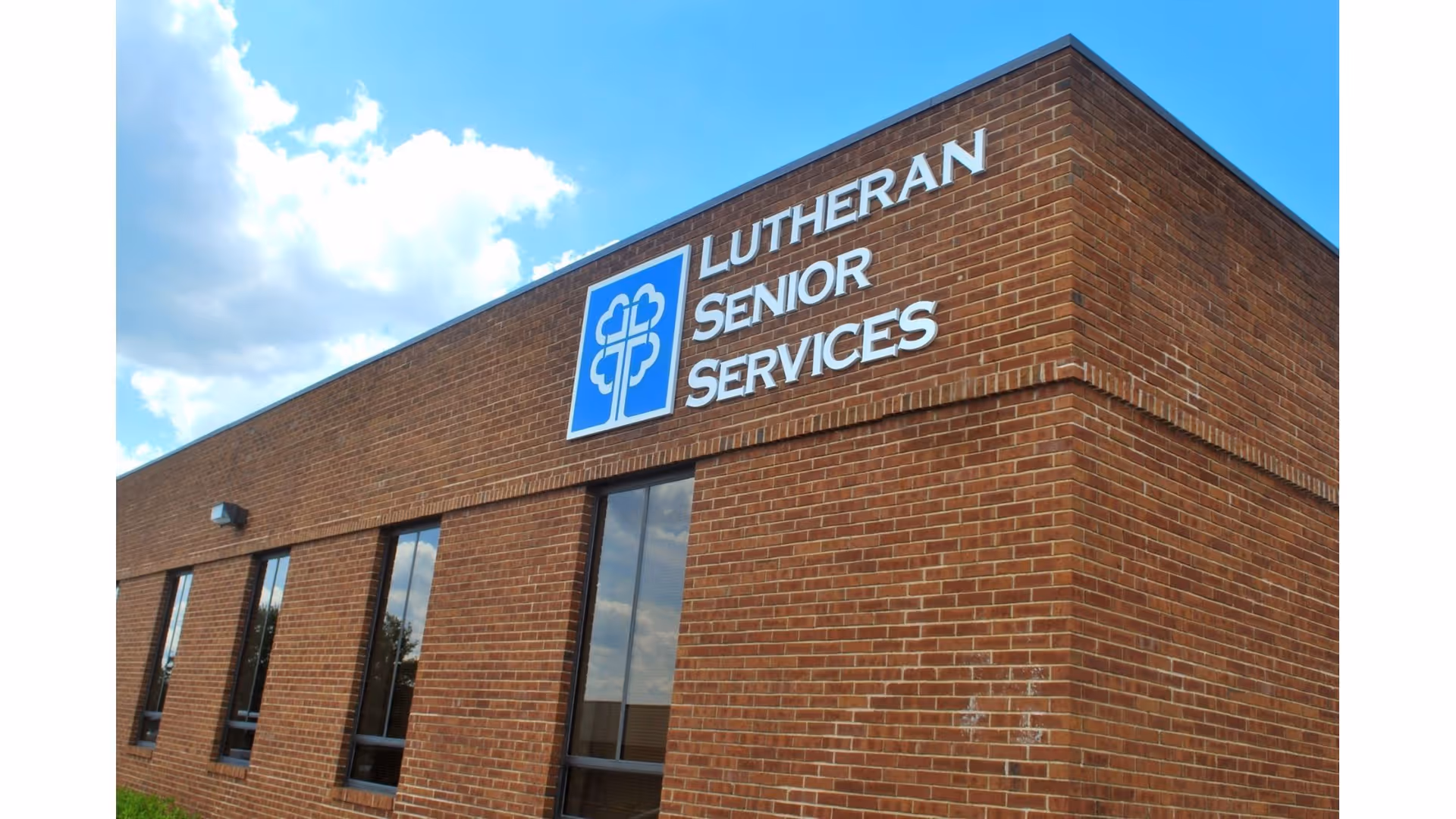 Exterior view of a brick building with a sign that reads 'Lutheran Senior Services' under a blue sky with some clouds.