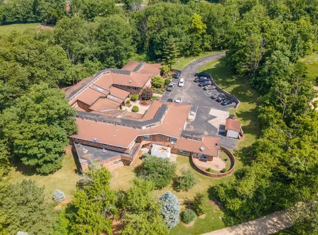 Aerial view of Brookwood Retirement Community showing a large building with a red roof surrounded by dense green trees and a parking lot with several cars.