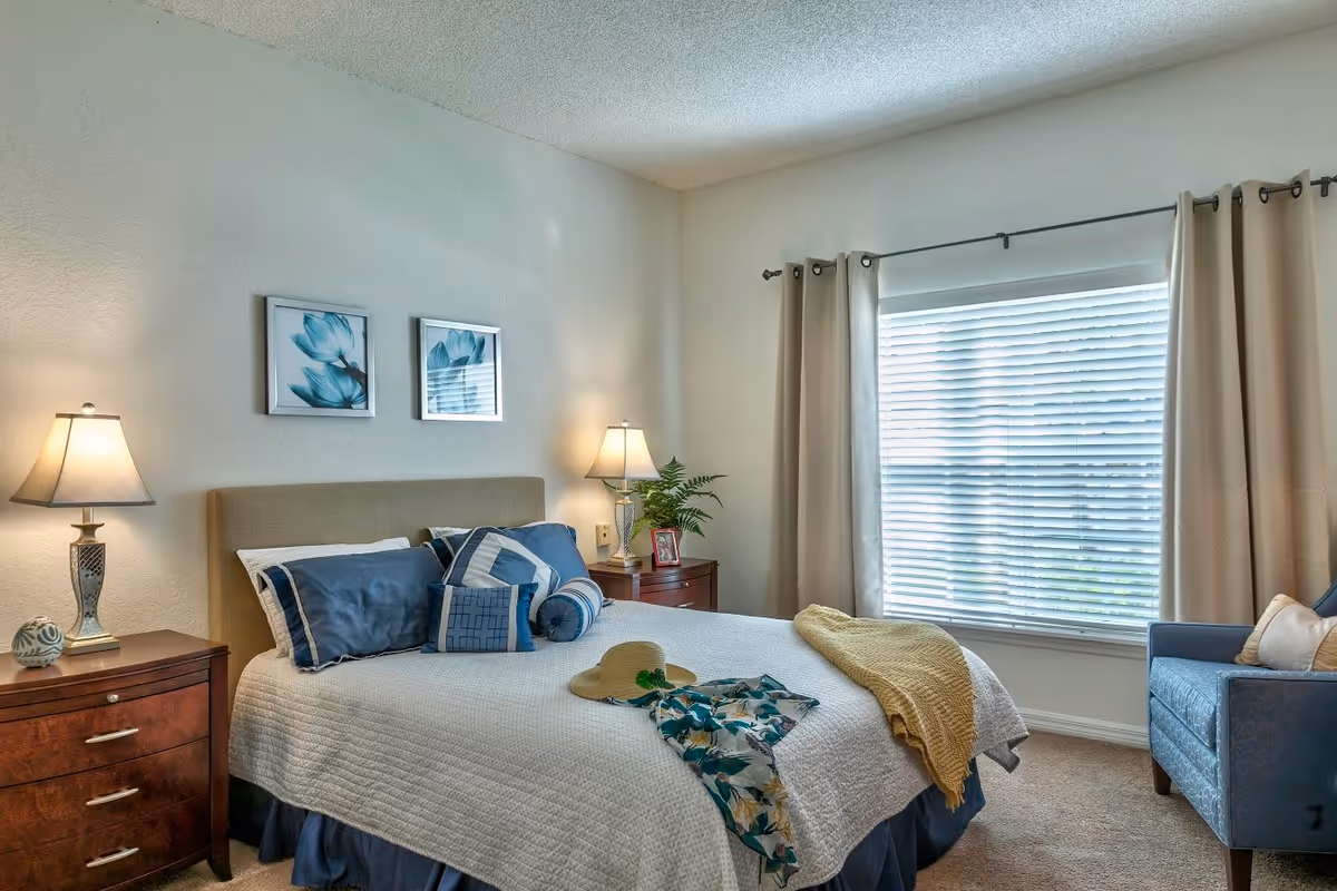 A cozy bedroom with a neatly made bed featuring blue and white bedding, two bedside tables with lamps, framed floral artwork on the wall, a large window with beige curtains, and a blue armchair in the corner. A yellow throw blanket, a sun hat, and a floral garment are placed on the bed.