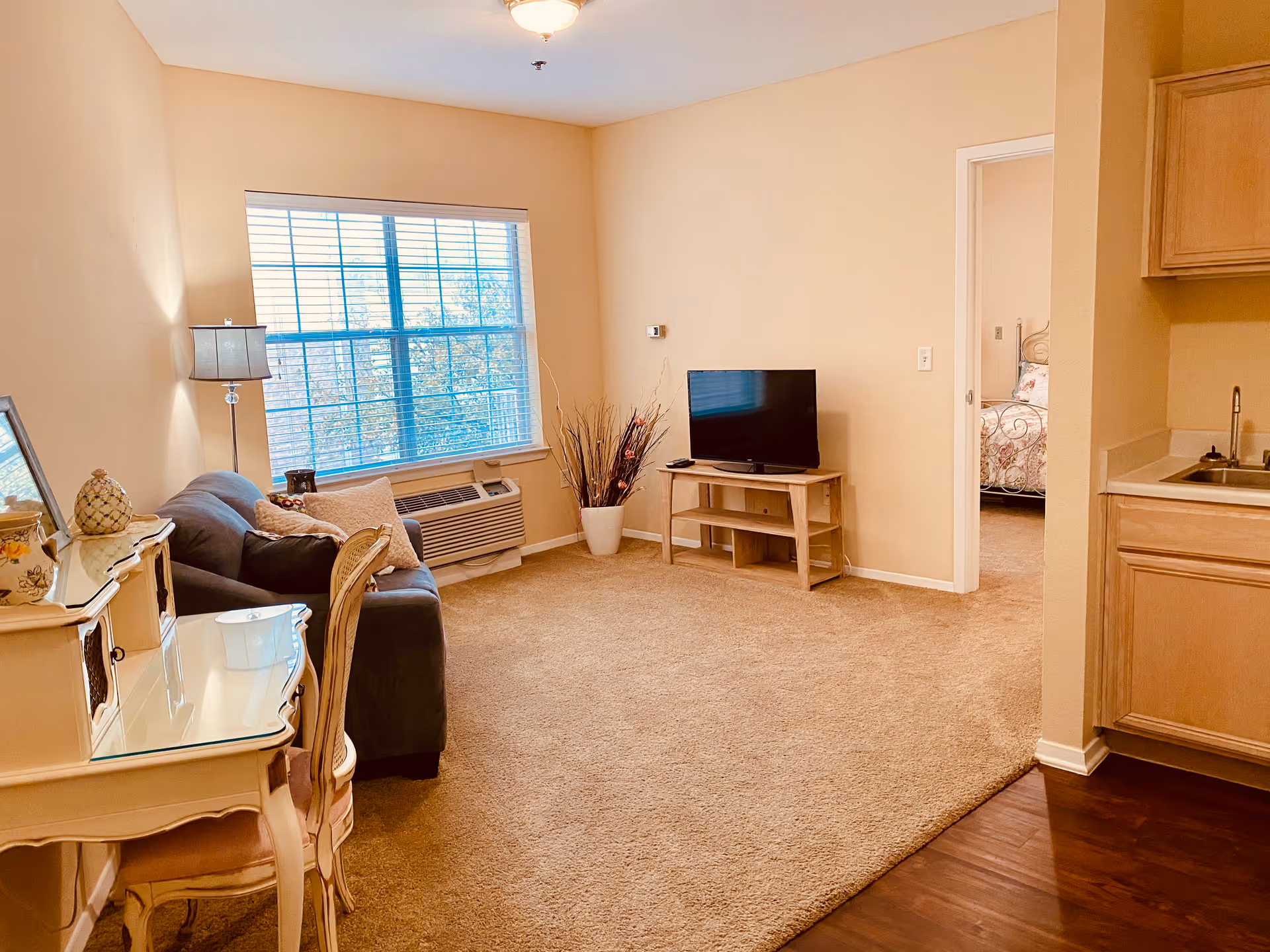 A cozy living room with beige walls and carpet, featuring a dark gray sofa with pillows, a floor lamp, a small wooden TV stand with a flat-screen TV, and a window with blinds letting in natural light. To the right, there is a partial view of a kitchen area with wooden cabinets and a sink, and an open doorway leading to a bedroom with a floral bedspread.