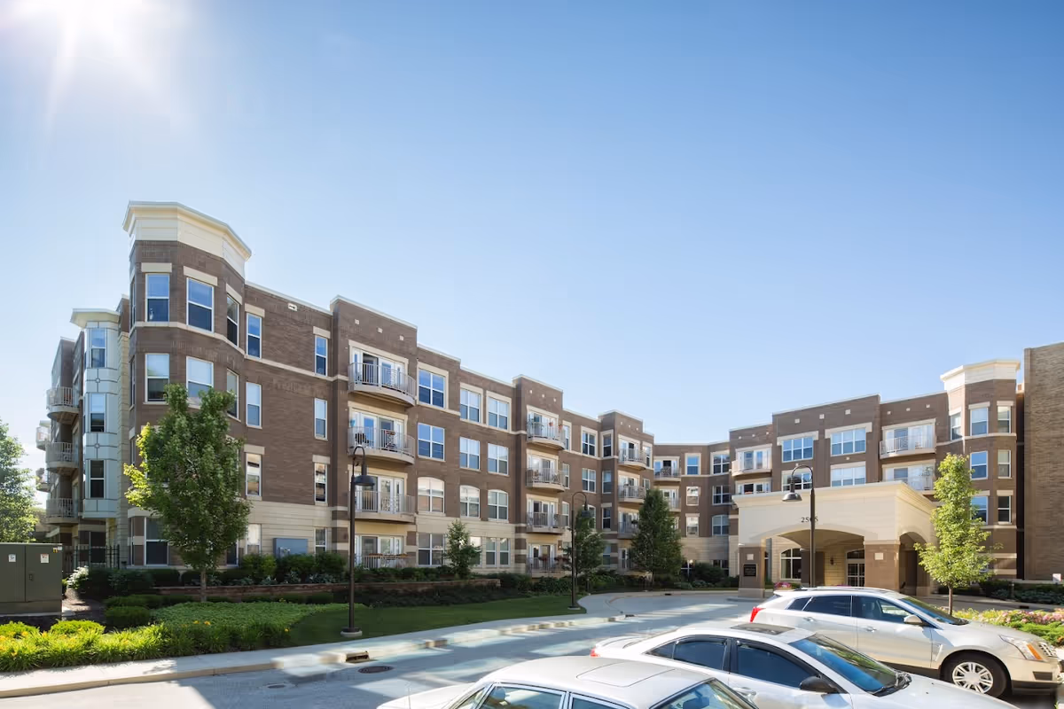Exterior view of a multi-story senior living facility named Eastcastle Place with a brick facade, multiple windows, balconies, and a covered entrance. Several cars are parked in front of the building, and there are trees and landscaped greenery around the property under a clear blue sky.