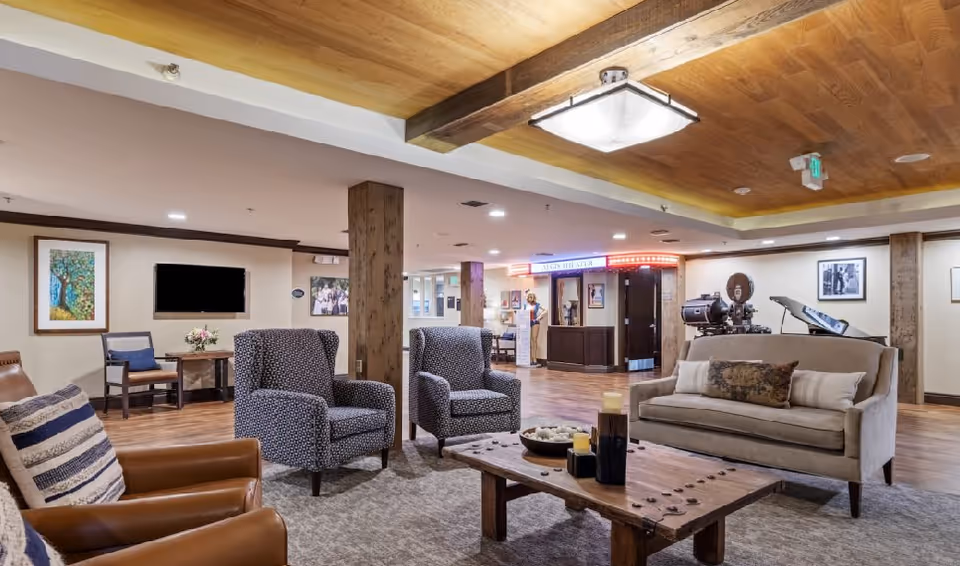 A cozy senior living facility common area with comfortable seating including two patterned armchairs, a beige sofa with pillows, and a leather chair with a striped pillow. A wooden coffee table with decorative candles and a bowl is in the center. The room has wooden ceiling beams, hardwood floors, framed artwork on the walls, and a reception desk in the background with a neon sign reading 'Aegis Theater'.