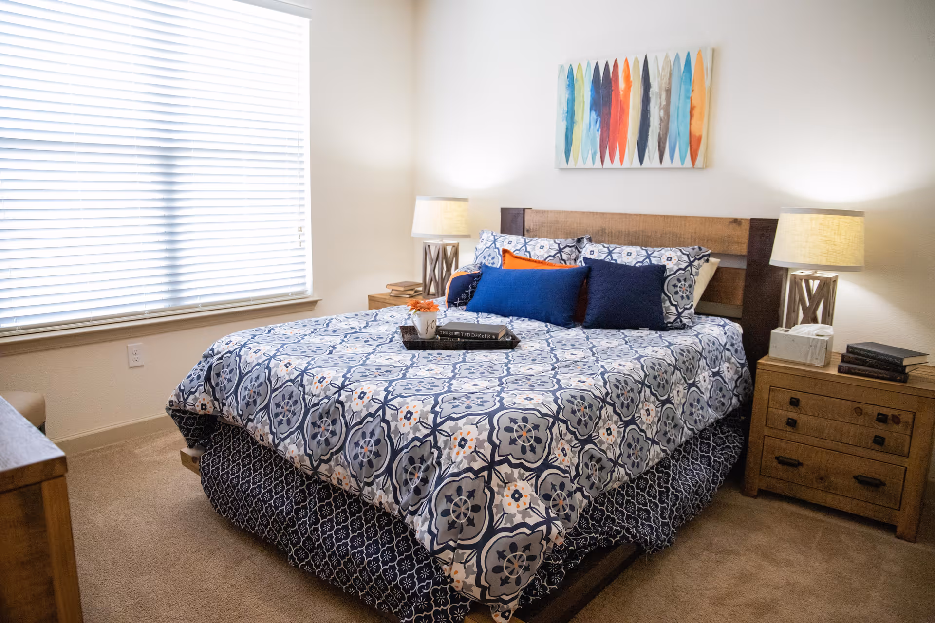 A cozy bedroom with a bed covered in a blue and white patterned comforter and multiple pillows. There are two wooden nightstands on either side of the bed, each with a lamp. A colorful abstract painting hangs above the bed, and a window with closed blinds is on the left side of the room.