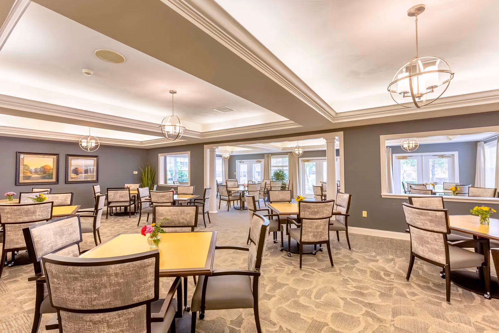 A spacious dining room with multiple square tables and cushioned chairs arranged neatly. The room features soft lighting from modern pendant lights, large windows allowing natural light, framed artwork on the walls, and small flower arrangements on each table. The carpet has a subtle floral pattern, and the walls are painted in a muted gray tone.