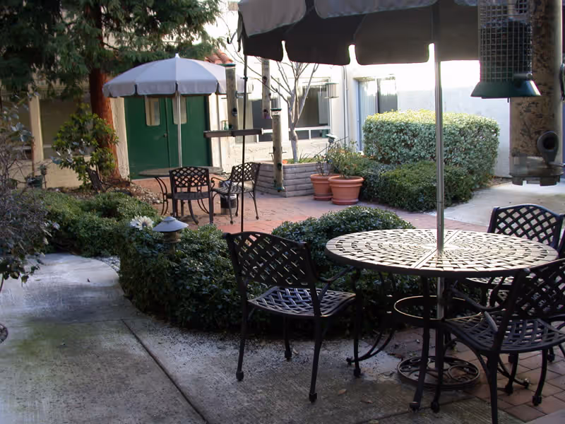 Outdoor patio area with metal tables and chairs, some with umbrellas. Surrounding the patio are green bushes, potted plants, and a concrete walkway. A building with green double doors and windows is visible in the background.