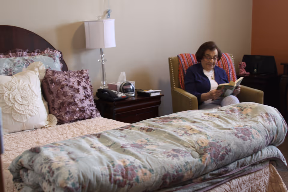A cozy bedroom with a neatly made bed featuring floral bedding and decorative pillows. Next to the bed is a nightstand with a lamp, phone, and clock. An elderly woman is sitting in an armchair reading a book, with a colorful blanket draped over the back of the chair.