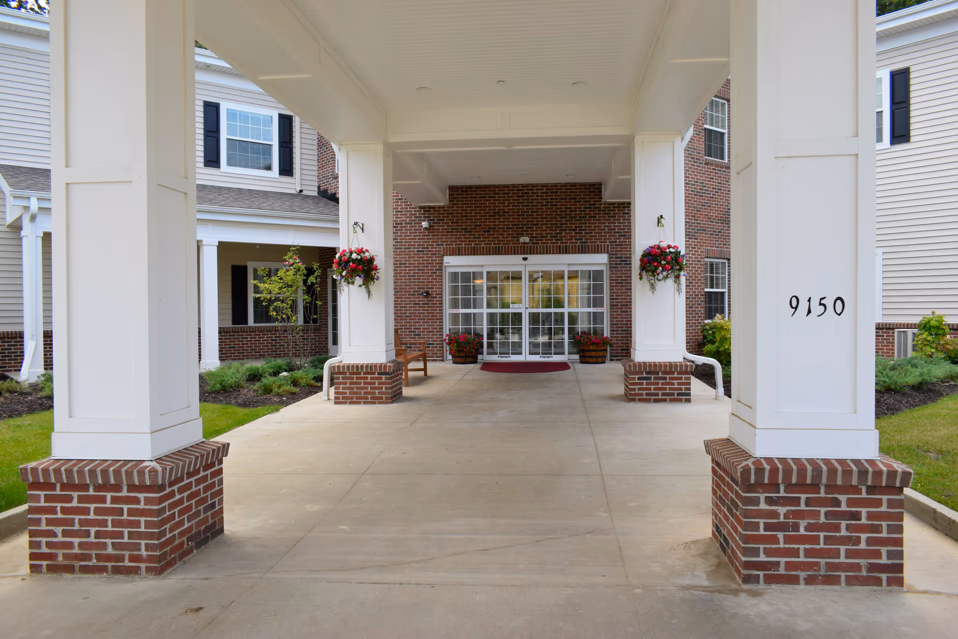 Covered entrance to a senior living facility with white pillars on brick bases, hanging flower baskets, and automatic glass doors. The building exterior features brick and siding with black shutters on the windows. The number 9150 is displayed on one of the pillars.
