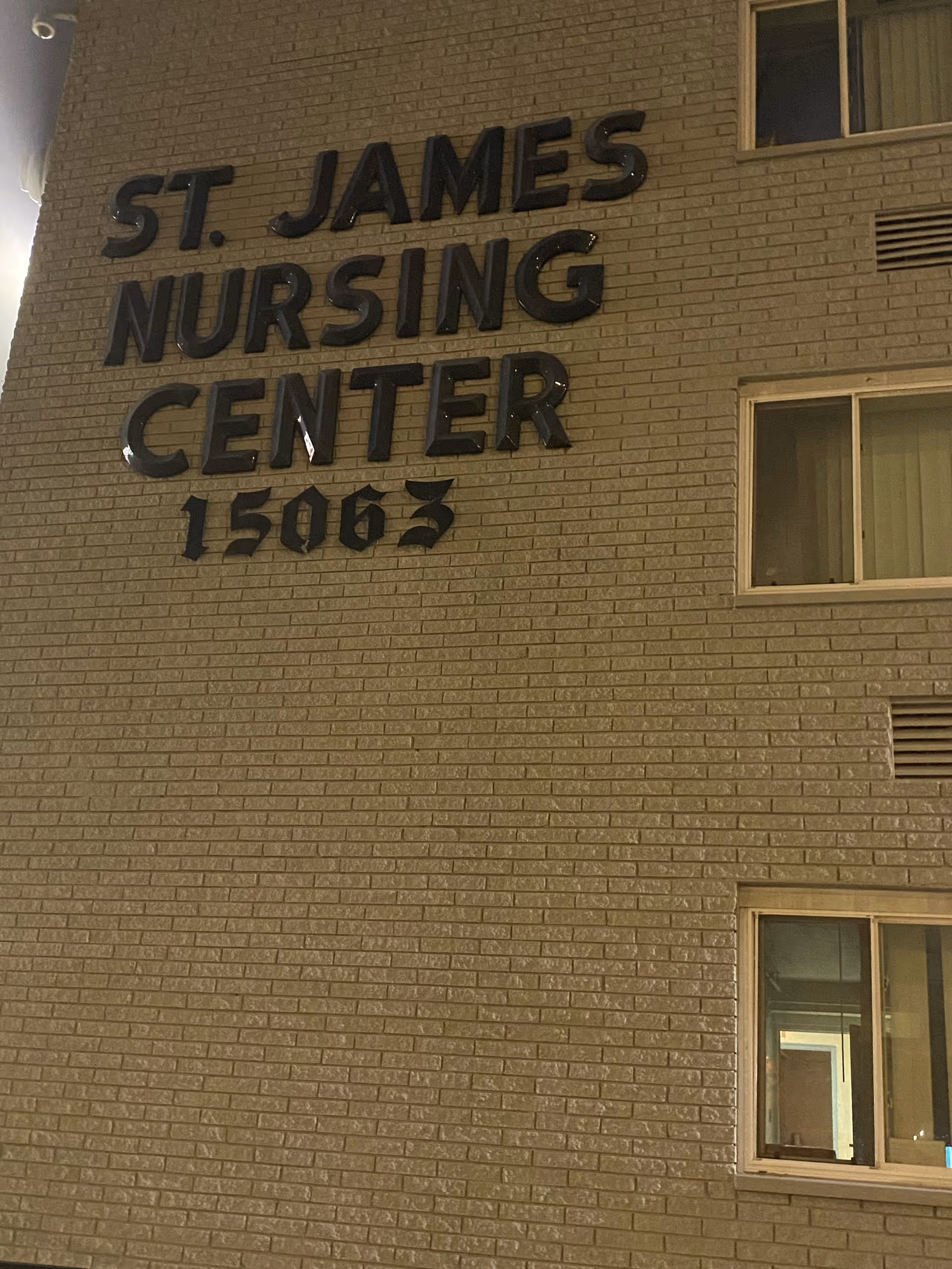 Exterior wall of a building with the sign 'ST. JAMES NURSING CENTER 15063' mounted on the brick surface, along with three windows visible on the right side.