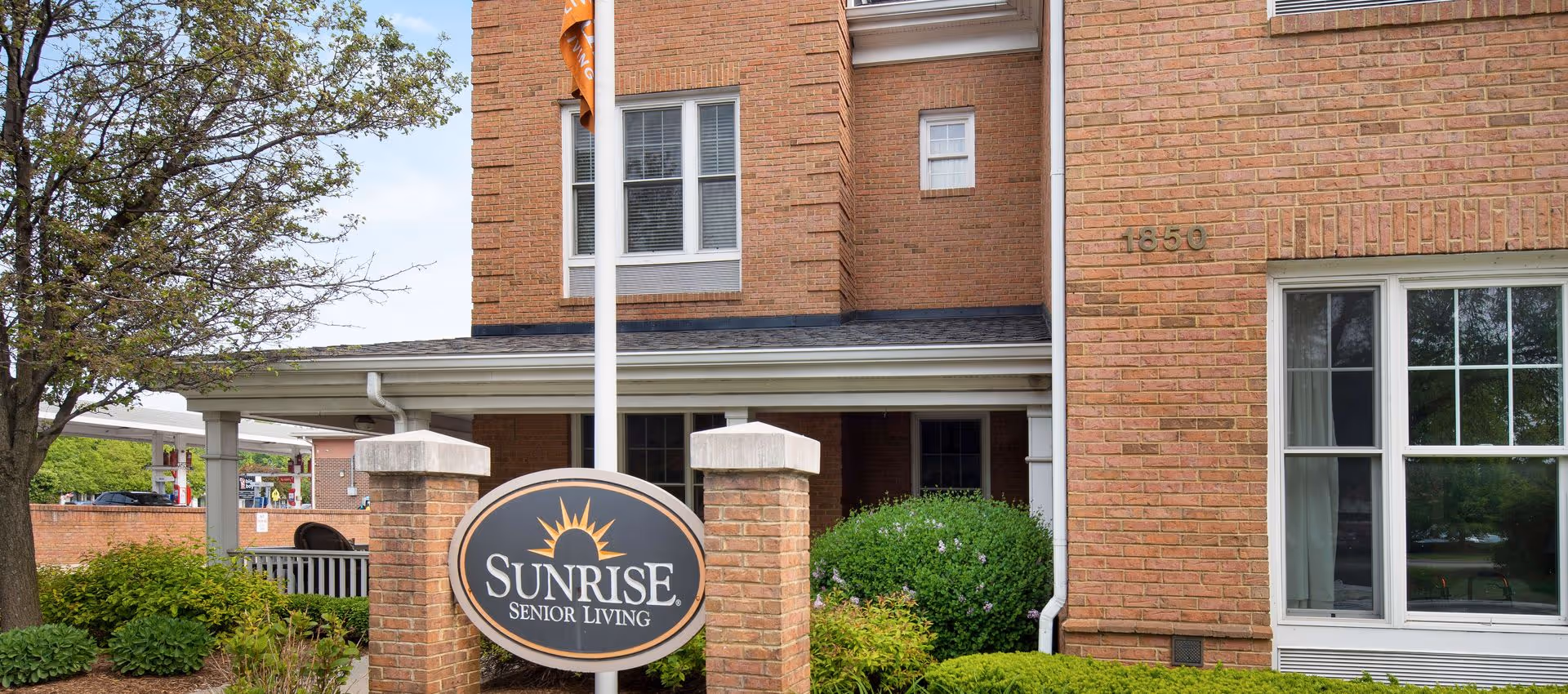 Exterior view of a brick building with a sign in front that reads 'Sunrise Senior Living'. The building has multiple windows, a covered entrance, and is surrounded by green bushes and a tree. The address number 1850 is visible on the building.