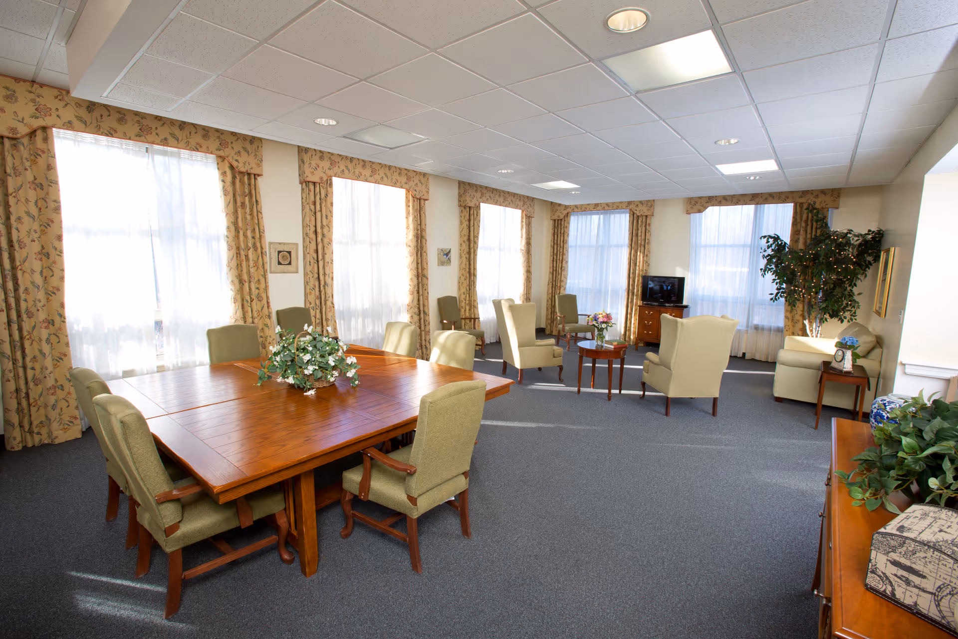 A spacious, well-lit common area with large windows covered by floral curtains. The room features a wooden table surrounded by green upholstered chairs, a seating area with armchairs and small tables, a TV on a wooden stand, and decorative plants and flowers throughout.