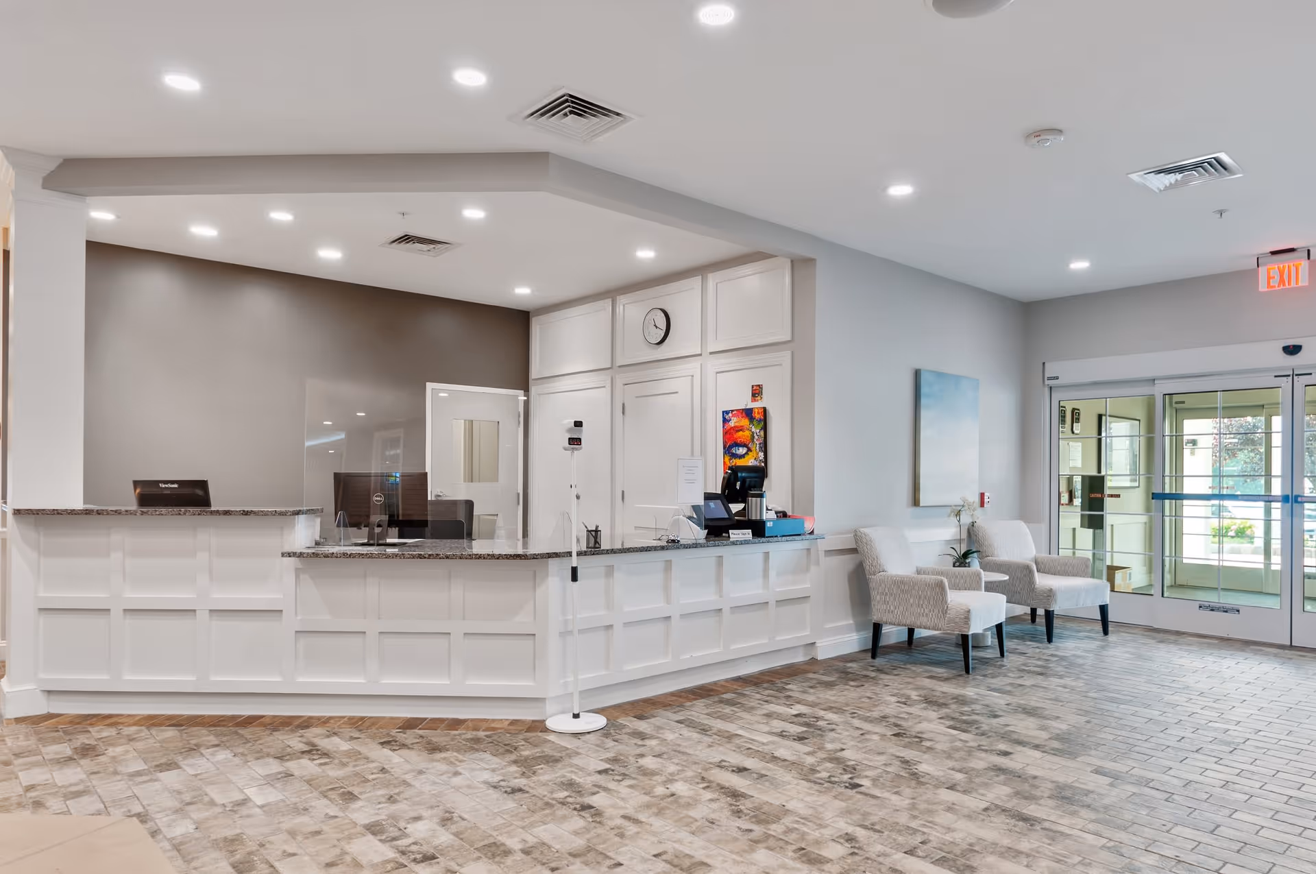 Reception area of a senior living facility with a white paneled front desk, two beige armchairs with a small table and plant between them, a clock on the wall, and glass automatic entrance doors letting in natural light.