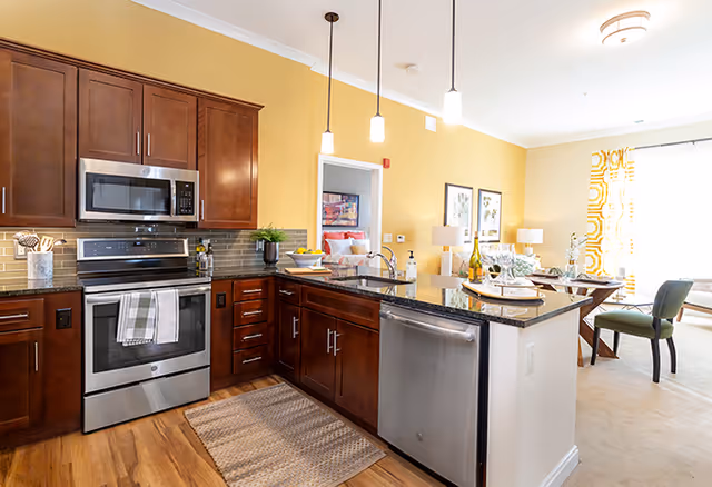 A modern kitchen with dark wood cabinets, stainless steel appliances including a microwave, oven, and dishwasher. The kitchen has a granite countertop island with a sink and three pendant lights hanging above. The background shows a dining area with a table, chairs, and bright yellow patterned curtains on the window.