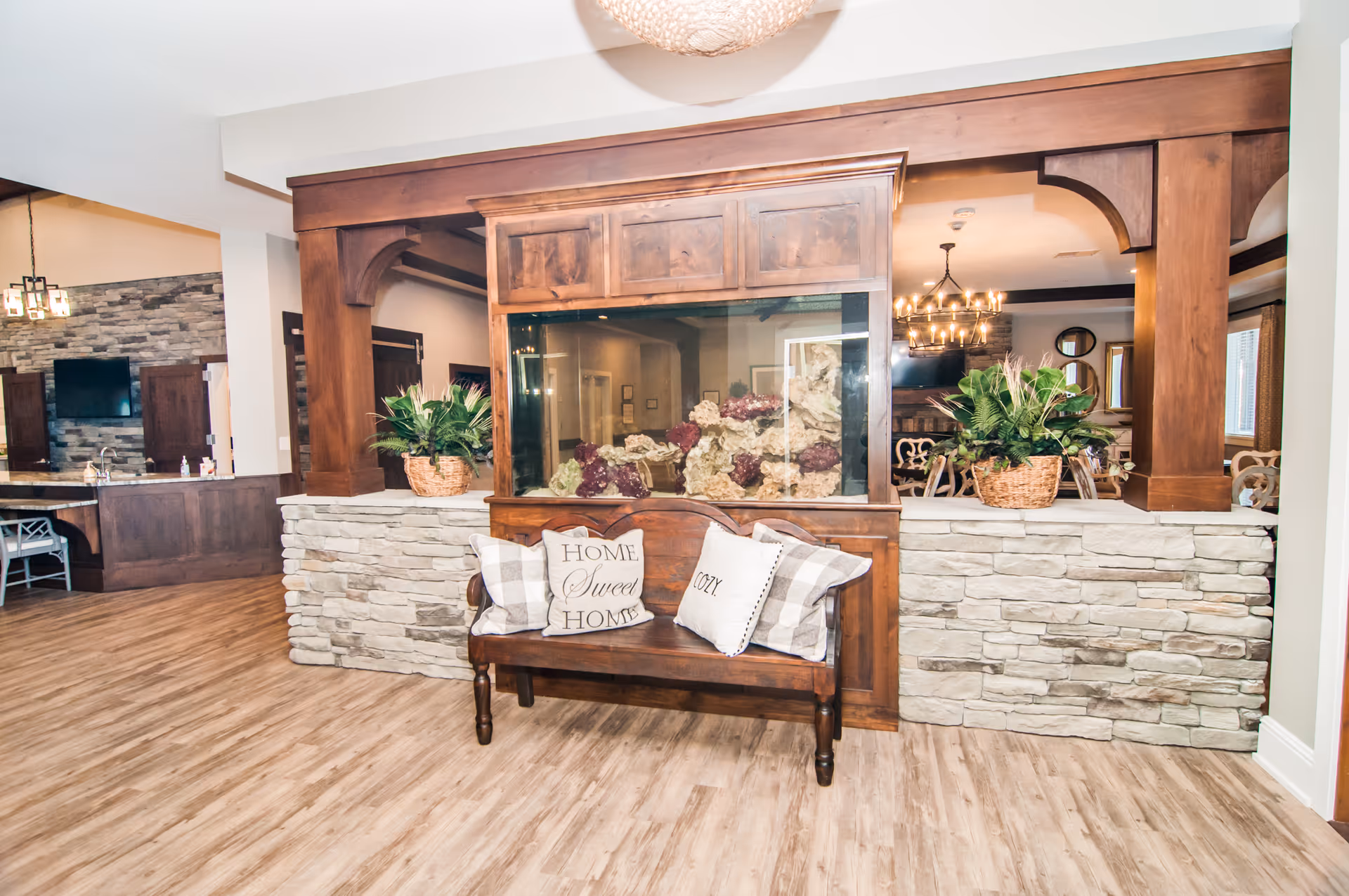 Interior view of a senior living facility common area featuring a wooden bench with decorative pillows that say 'Home Sweet Home' and 'Cozy'. Behind the bench is a large built-in aquarium with rocks and plants, flanked by stone half-walls and wooden columns. The background shows a dining area with a chandelier and a kitchen area with a stone backsplash and a TV mounted on the wall.