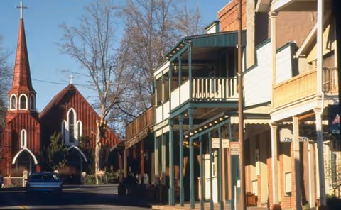 Street view of a small town with a red church featuring a tall steeple on the left side and a row of buildings with balconies and awnings on the right side under a clear blue sky.