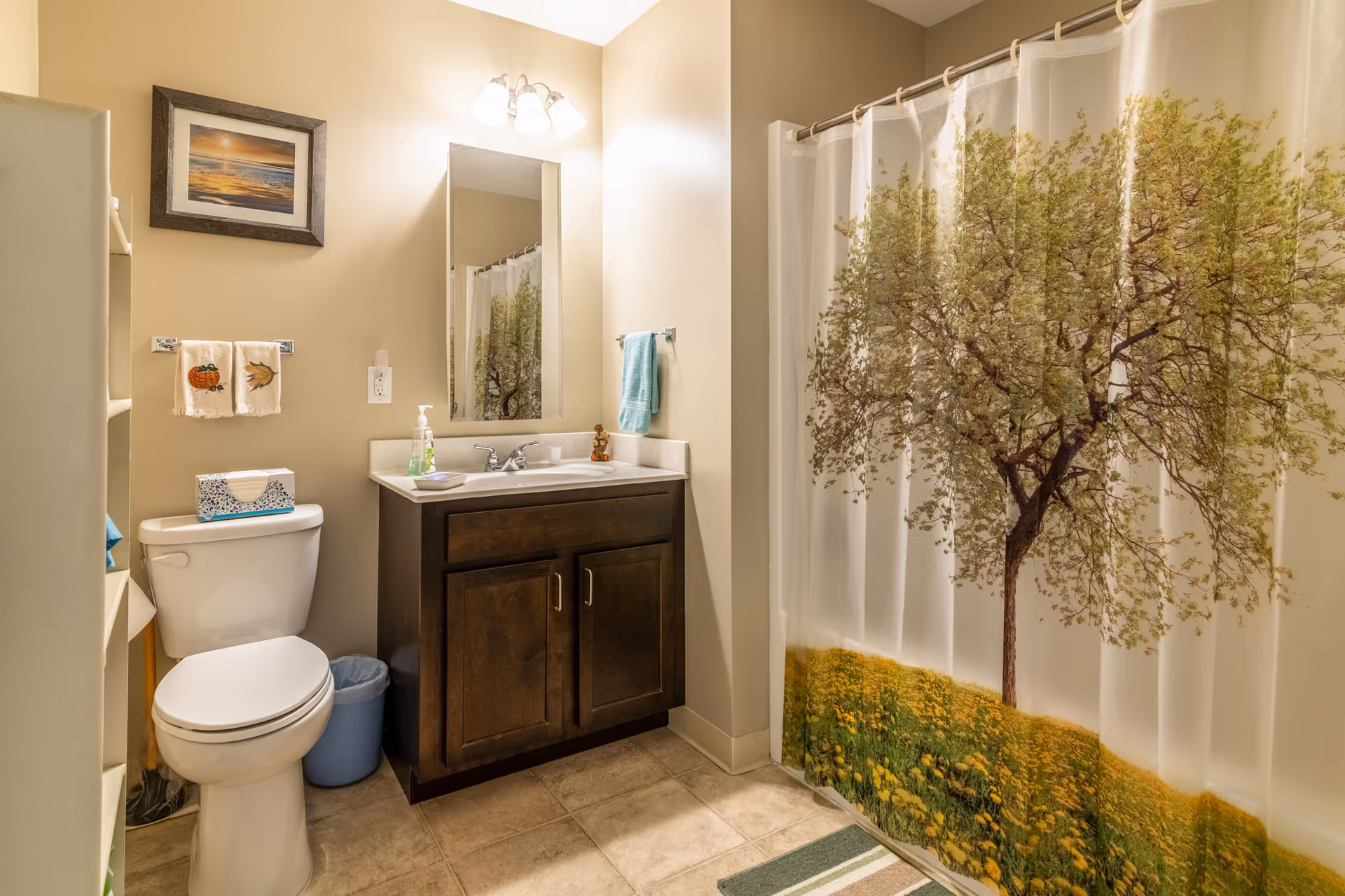 A bathroom with beige walls and tiled floor featuring a white toilet, a dark wood vanity with a sink and mirror above it, a shower with a curtain depicting a tree and a field of yellow flowers, a small blue trash bin, and decorative towels hanging on the wall.