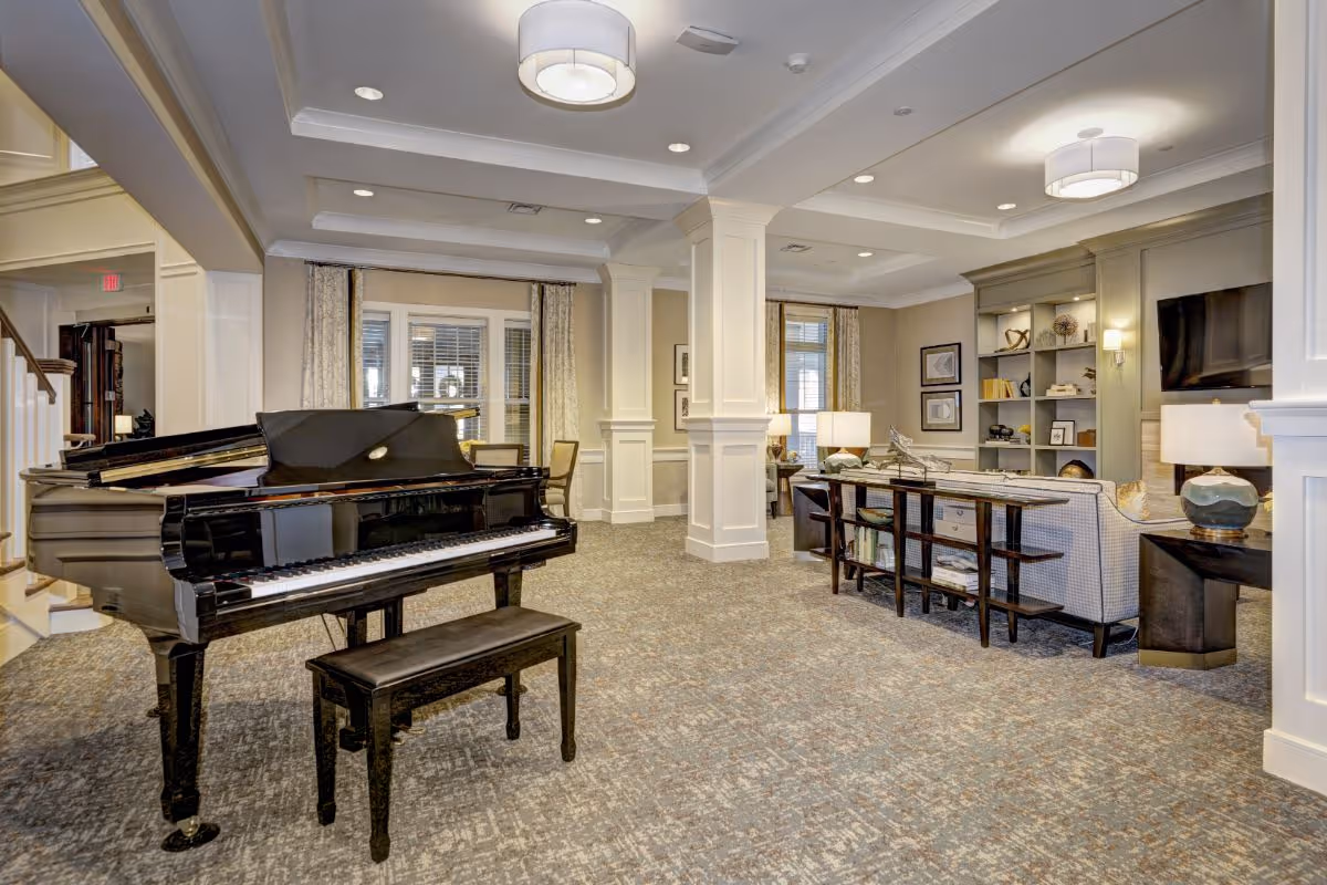 Spacious and well-lit living room area in a senior living facility featuring a black grand piano with a matching bench, beige carpeted floor, white pillars, a seating area with a sofa and side tables with lamps, built-in shelves with decorative items, and a wall-mounted flat-screen TV.