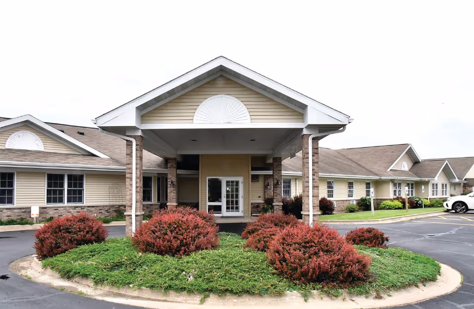 Front exterior view of a single-story senior living facility building with beige siding, brick accents, and a covered entrance. There are red bushes and green shrubs in a landscaped area in front of the entrance, and a parking lot surrounds the building.