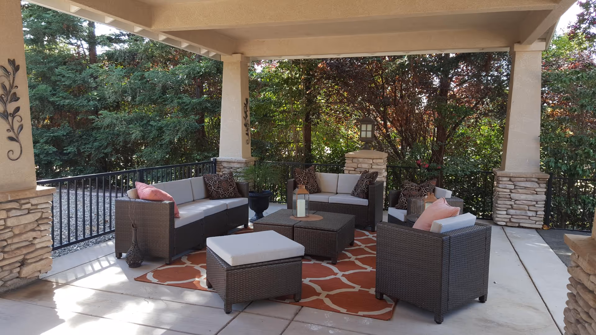 Covered outdoor patio area with cushioned wicker furniture including two sofas, two armchairs, and an ottoman arranged around a central coffee table on an orange patterned rug. The patio is surrounded by a black metal railing and lush green trees and shrubs in the background.