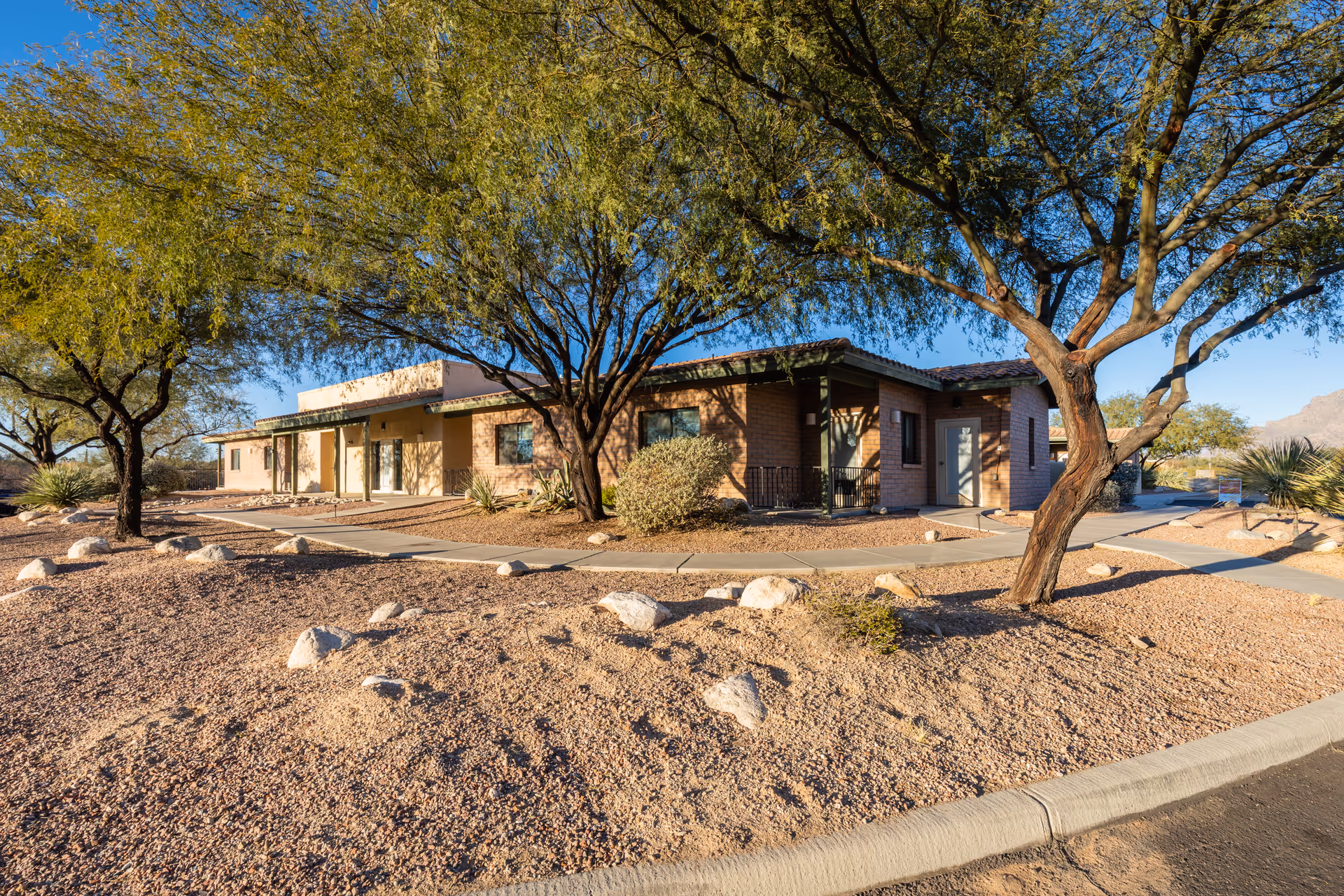Exterior view of a single-story assisted living facility building surrounded by desert landscaping with gravel, rocks, and several trees under a clear blue sky.