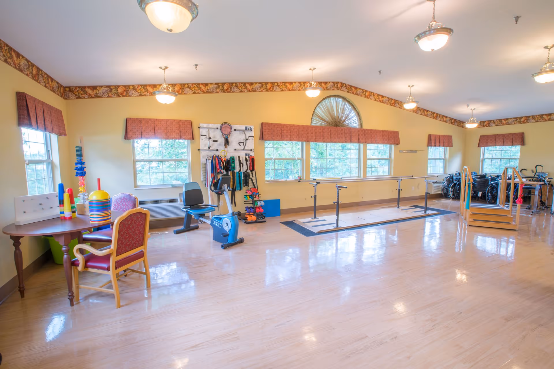 A spacious rehabilitation room with wooden floors and yellow walls, featuring exercise equipment including a stationary bike, parallel bars for walking practice, and various therapy tools on a pegboard. There are several windows with red valances letting in natural light, a small table with chairs, and a set of stairs with handrails for physical therapy exercises.
