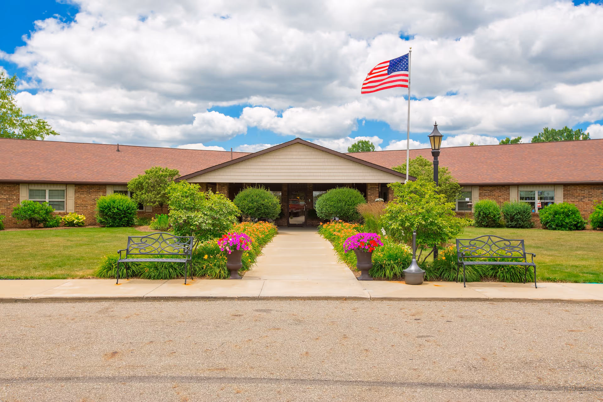Front exterior view of Canterbury Villa of Alliance, a single-story brick building with a brown roof. The entrance is centered with a walkway flanked by green bushes and colorful flower pots. Two black metal benches are placed on either side of the walkway. An American flag on a flagpole and a vintage-style street lamp are visible near the entrance. The sky is partly cloudy with patches of blue.