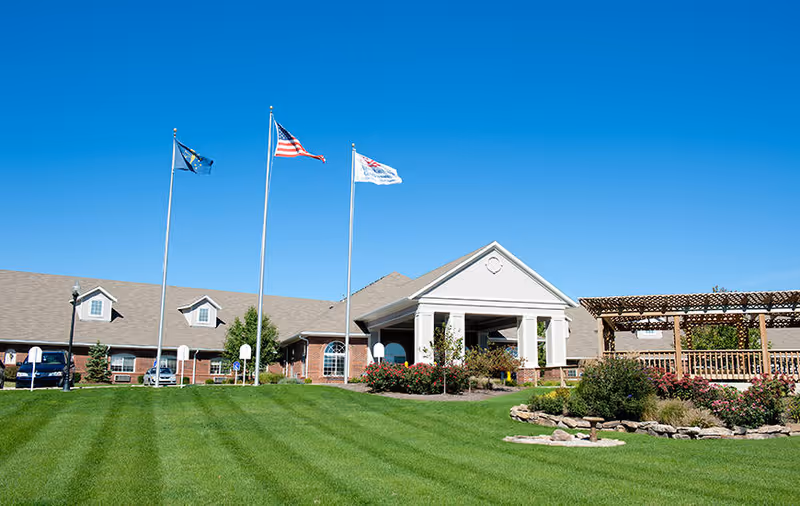 Exterior view of Countryside Meadows senior living facility with a well-maintained green lawn, three flagpoles flying flags, a brick building with a covered entrance, and a wooden pergola on the right side under a clear blue sky.