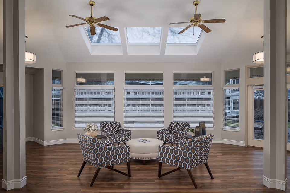 A bright and spacious sitting area with four patterned armchairs arranged around a round ottoman in the center. The room features large windows with blinds, three skylights on the ceiling, two ceiling fans, and wood flooring. The walls are painted light gray, and there are two small side tables with decorative items.