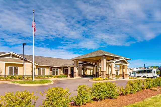 Exterior view of Arbor Oaks Terrace Memory Care Residence showing a single-story building with a covered entrance, an American flag on a flagpole, a driveway, landscaped bushes, and a shuttle bus parked on the right side under a partly cloudy blue sky.