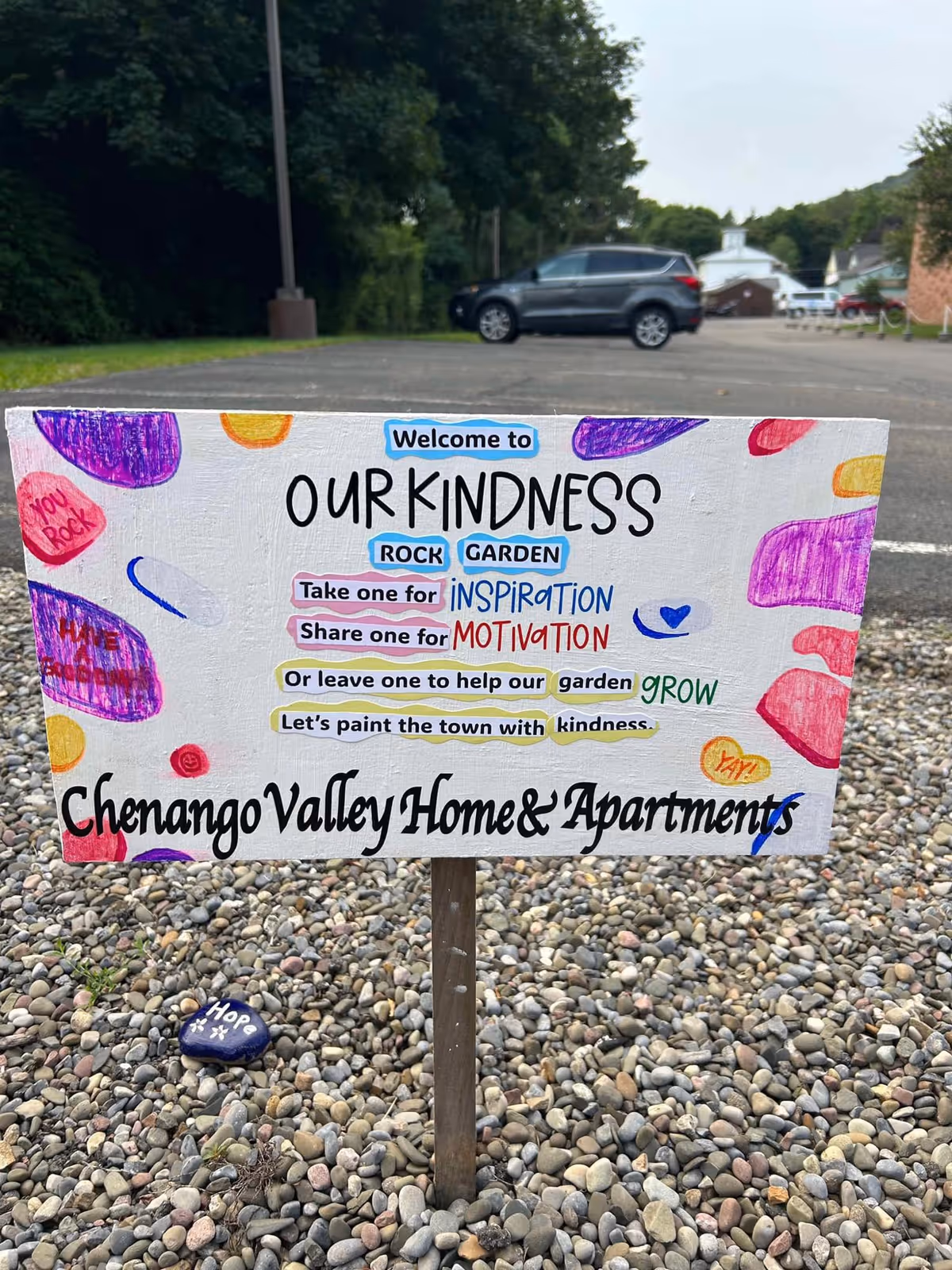 A colorful hand-painted sign in a rock garden with pebbles and a painted rock that says 'Hope'. The sign reads: 'Welcome to Our Kindness Rock Garden. Take one for Inspiration. Share one for Motivation. Or leave one to help our garden grow. Let's paint the town with kindness. Chenango Valley Home & Apartments.' The background shows a parking lot with a parked car and trees.