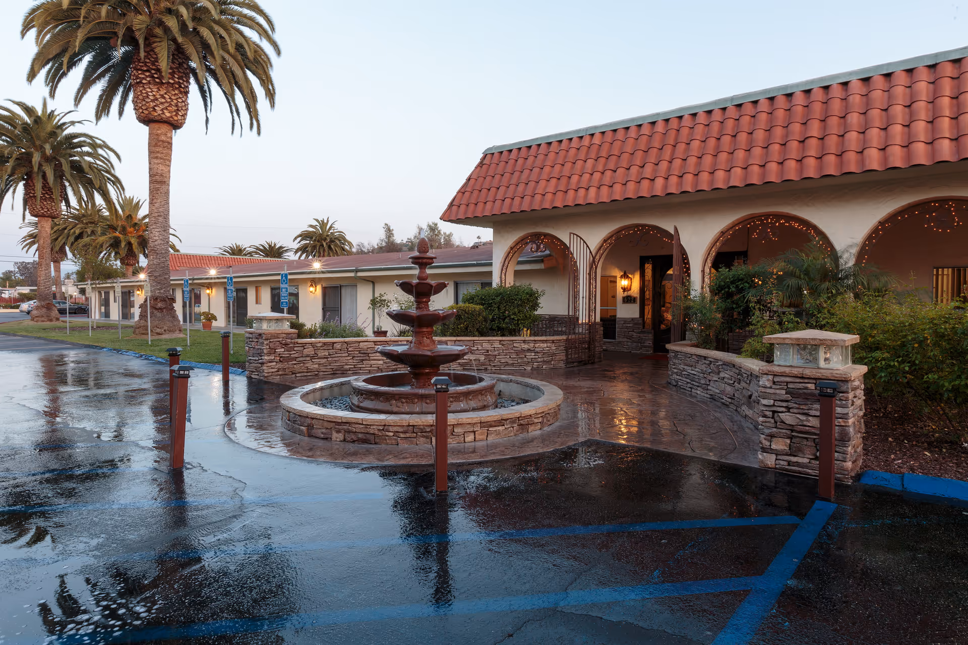 Exterior view of Villa Las Palmas Healthcare Center featuring a wet driveway with blue handicap parking spaces, a multi-tiered stone fountain in front of the entrance, palm trees, and a building with a red tiled roof and arched doorways decorated with string lights.