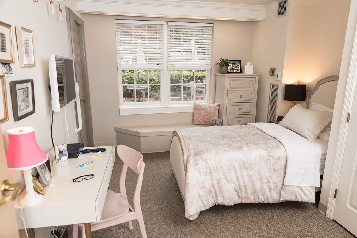 A cozy bedroom with a single bed covered in a light pink and beige comforter. There is a beige headboard, a nightstand with a black lamp, and a tall dresser with a plant and framed photos on top. A window with white blinds overlooks greenery outside. A white desk with a pink lamp, glasses, and a chair is positioned against the wall, along with a wall-mounted TV and several framed pictures.