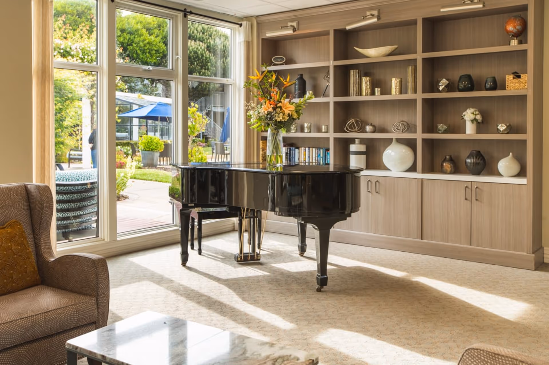 A bright living room area with a black grand piano adorned with a vase of flowers. Behind the piano is a large built-in wooden bookshelf filled with decorative items and books. Large windows reveal a view of a garden patio with outdoor seating and umbrellas.
