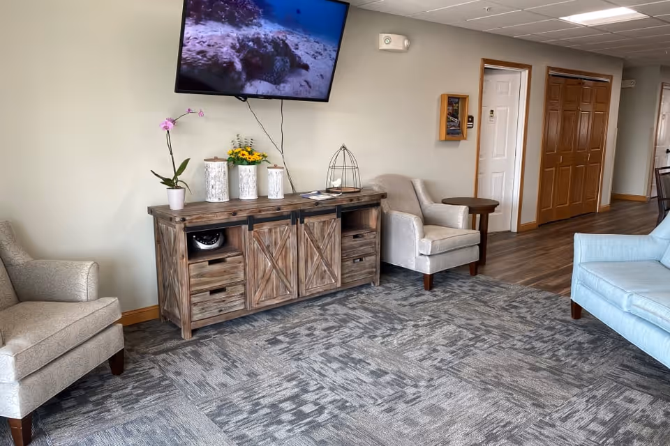 A cozy living room area with a patterned carpet, two beige armchairs, a light blue sofa, and a rustic wooden cabinet with sliding barn doors. A flat-screen TV is mounted on the wall above the cabinet, displaying an underwater scene. The room has light-colored walls, wooden trim, and a hallway with wooden doors in the background.