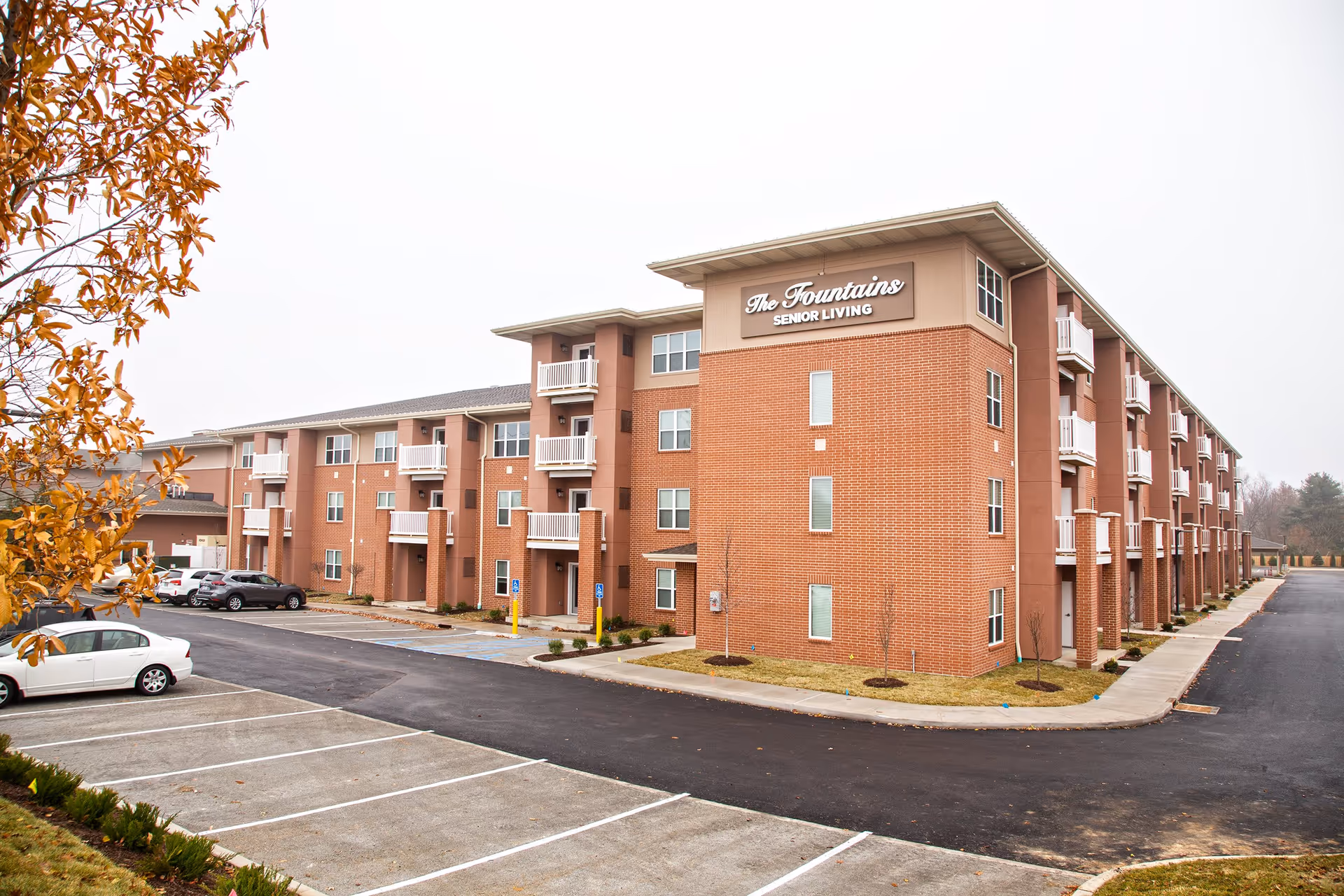 Exterior view of The Fountains Senior Living facility, a three-story brick building with multiple balconies and a parking lot in front. The sky is overcast and there are some autumn-colored trees on the left side.