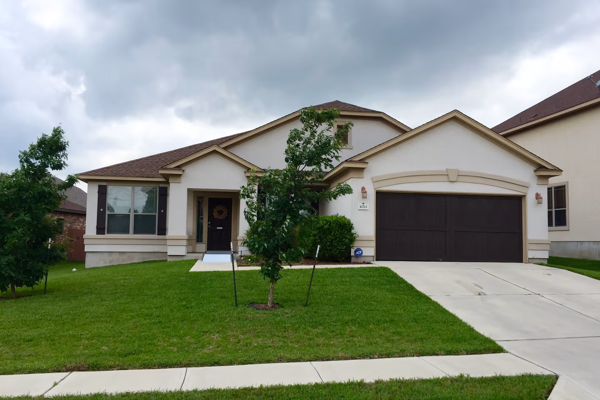 Front exterior view of a single-story house with a well-maintained green lawn, a concrete driveway leading to a double garage with dark brown doors, and a small tree in the front yard under a cloudy sky.